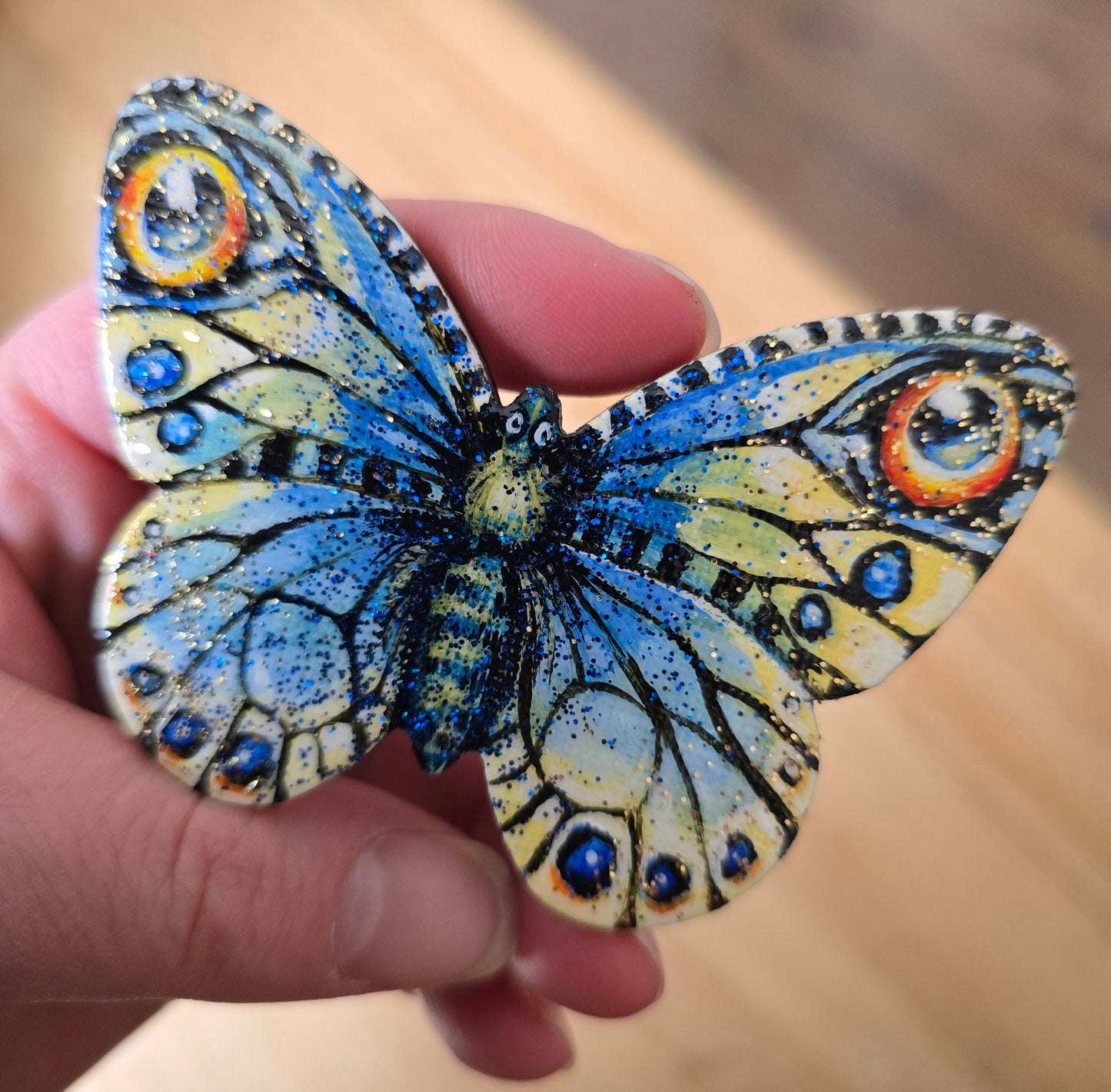 Sommerfugl i blått og gult med øyetegninger. Blått og gull glitter i den blanke fernissen Liten kjøleskapsmagnet håndmalt på sommerfuglfasonglerret.