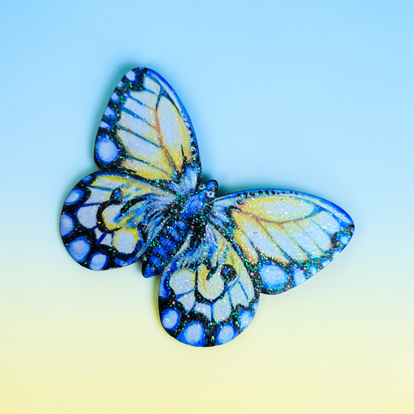 Liten håndmalt sommerfugl i blått og gule toner. med kult glitter i fernissen. Malt på lettet med magnetisk bakside. Brukt akrylmaling. Kjøleskapsmagnet.