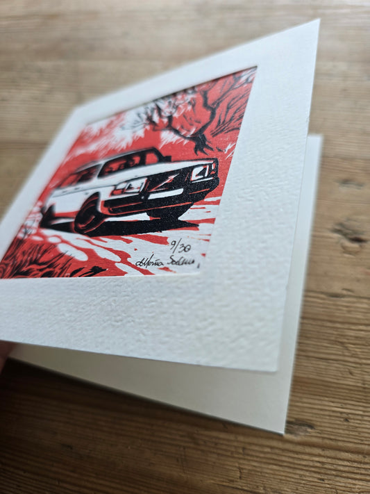 Linotrykk av Volvo 40 stasjonsogn sett forfra, Håndtrykket.med to plater så bildet har tre nyanser.  Hvitt som arket, rødt og svart. Bildet viser bilen og noe vegetasjon rundt. Trykket er montert i passepartoutkort med tilhørende konvolutt. Her blir kortet hold åpent for å vise at det er et dobbelt kort.