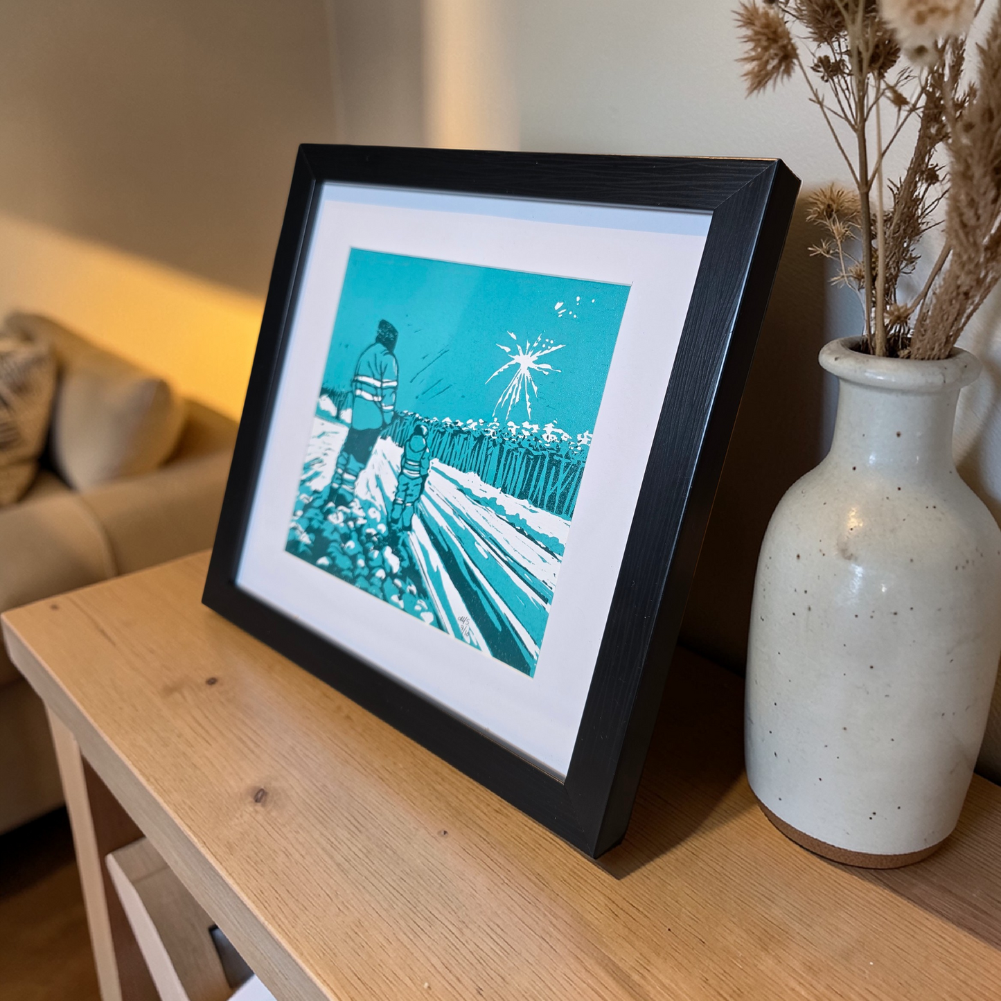 Unikt linotrykk, nummerert 7 av 10. Innrammet i svart ramme av kunststoff. Måler 22 x 22 cm. Motivet er av far og sønn som står sammen og ser nyttårsfyrverkeri, en hyggelig delt opplevelse. Fargene i bildet er mørke og lys grønnblå tone og hvitt i arket. Her er bildet sett fra skrått plassert på en hylle. Kunstner monica Sandmo