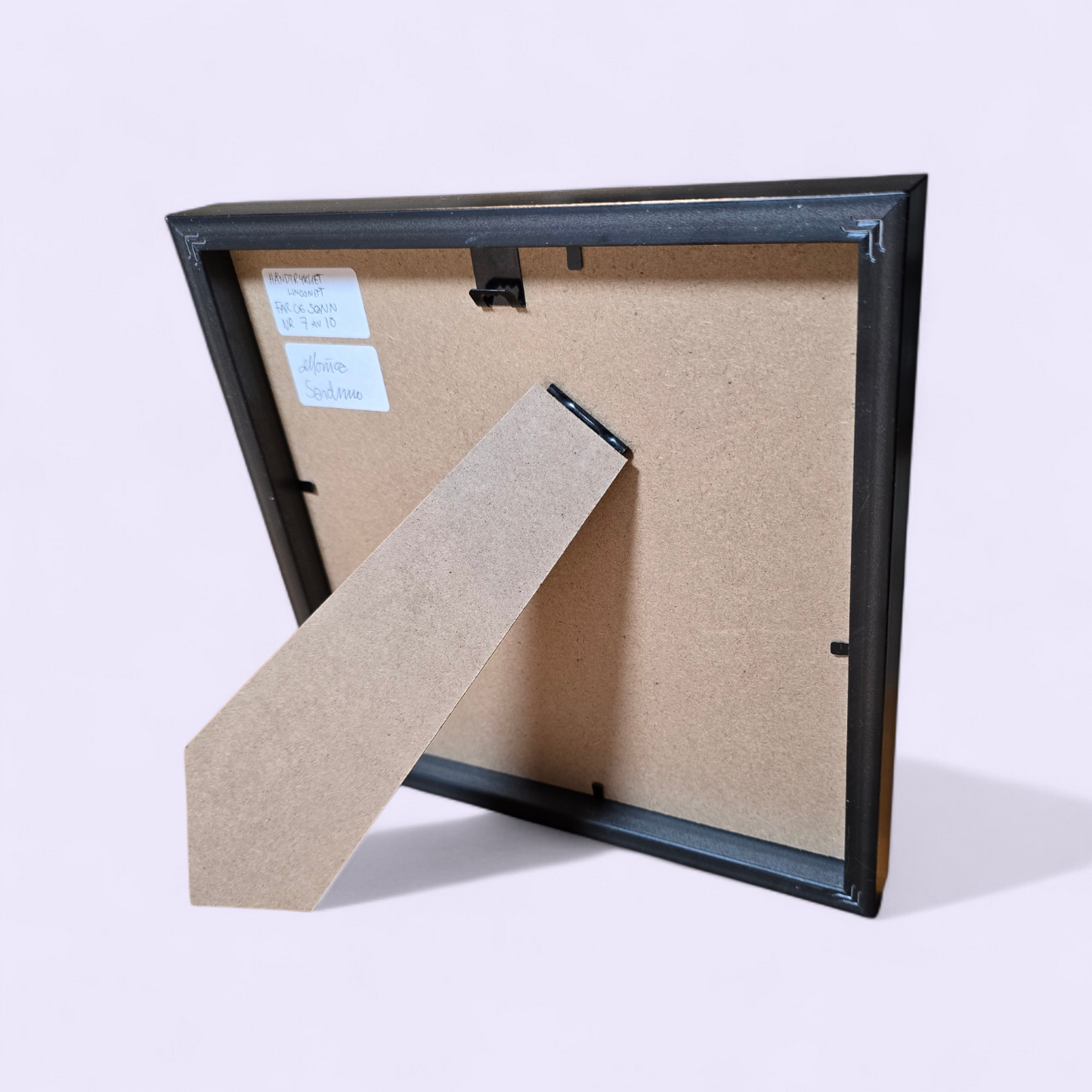 Unikt linotrykk, nummerert 7 av 10. Innrammet i svart ramme av kunststoff. Måler 22 x 22 cm. Motivet er av far og sønn som står sammen og ser nyttårsfyrverkeri, en hyggelig delt opplevelse. Fargene i bildet er mørke og lys grønnblå tone og hvitt i arket. Kunstner monica Sandmo
Dette er fotografi av baksiden av rammen bildet er montert inn i.