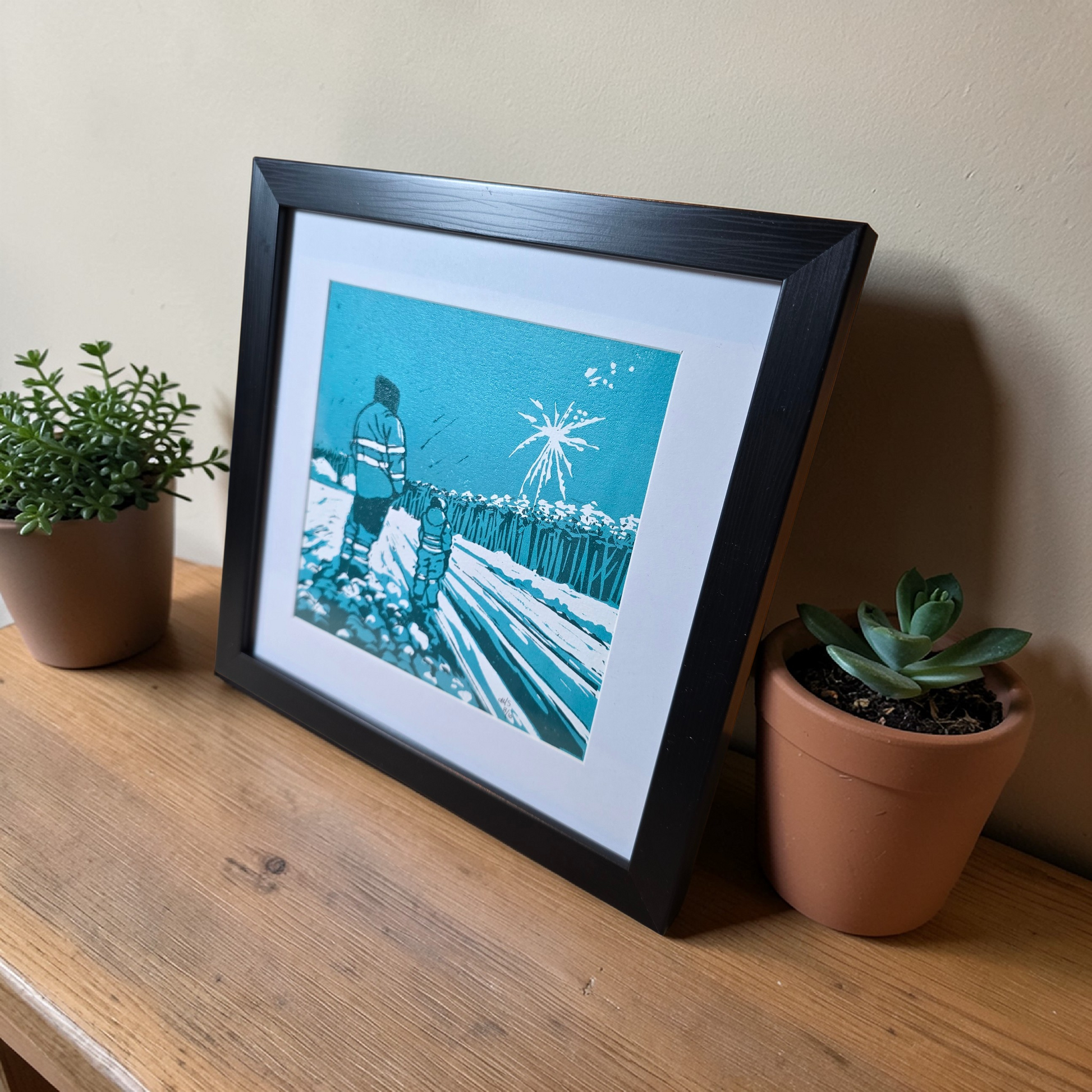 Linotrykk Far og sønn. Dette er nummerert  8 av 10.Trykket viser en far og sønn som står og ser nyttårsfyrverkeri sammen. En koselig stund. Trykket er trykket i blågrønt, mørk og lyst. så er det også hvitt fra arket. Hvit passeopartout og svart ramme i kunststoff. Her er bildet settfra siden , på en hylle med to planter.