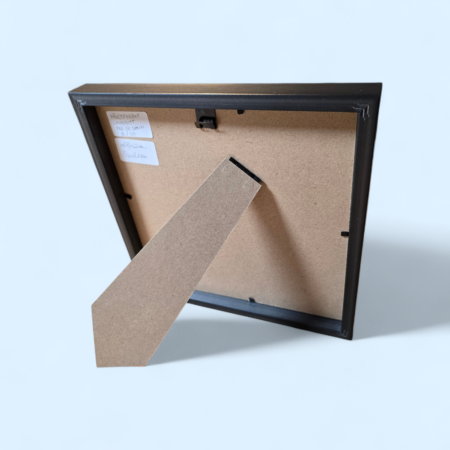 Linotrykk Far og sønn. Dette er nummerert  8 av 10.Trykket viser en far og sønn som står og ser nyttårsfyrverkeri sammen. En koselig stund. Trykket er trykket i blågrønt, mørk og lyst. så er det også hvitt fra arket. Hvit passeopartout og svart ramme i kunststoff.
Her er baksiden av bildet.