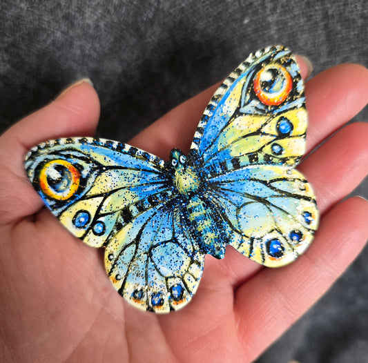 Sommerfugl i blått og gult med øyetegninger. Blått og gull glitter i den blanke fernissen Liten kjøleskapsmagnet håndmalt på sommerfuglfasonglerret.Her holdt i hånd.