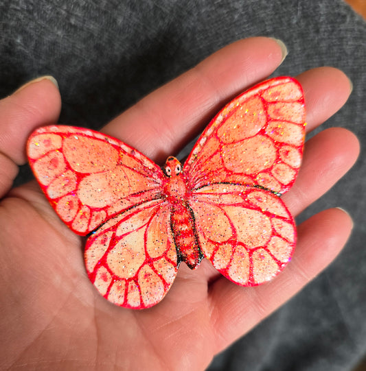 Liten håndmalt sommerfugl i rødt og fersken toner. med glitter i fernissen. Malt på lettet med magnetisk bakside. Brukt akrylmaling.Kjøleskapsmagnet.