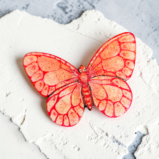 Liten håndmalt sommerfugl i rødt og fersken toner. med glitter i fernissen. Malt på lettet med magnetisk bakside. Brukt akrylmaling.Kjøleskapsmagnet.