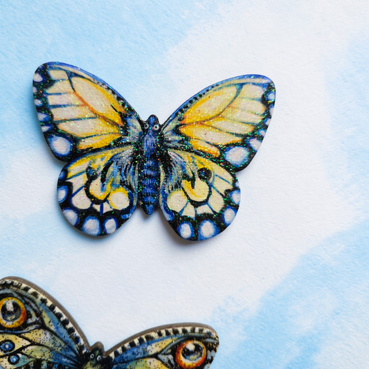 Liten håndmalt sommerfugl i blått og gule toner. med kult glitter i fernissen. Malt på lettet med magnetisk bakside. Brukt akrylmaling. Kjøleskapsmagnet.