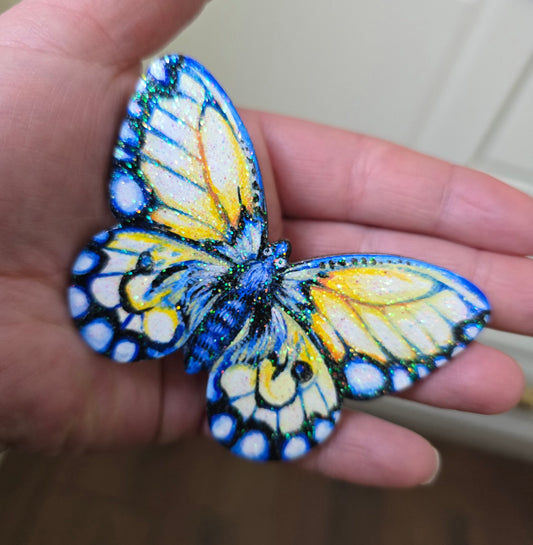 Liten håndmalt sommerfugl i blått og gule toner. med kult glitter i fernissen. Malt på lettet med magnetisk bakside. Brukt akrylmaling. Kjøleskapsmagnet.